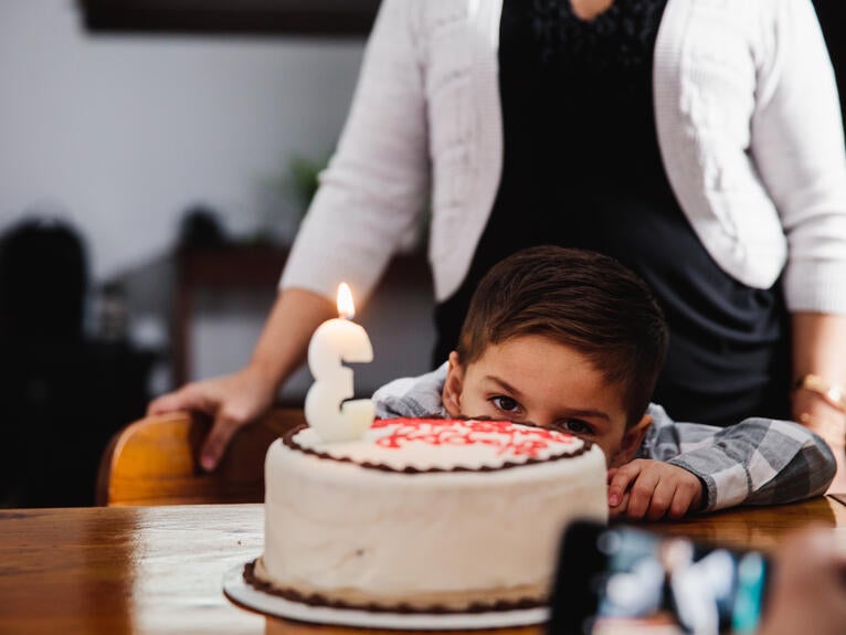 Boy hiding behind a birthday cake with an adult standing behind him